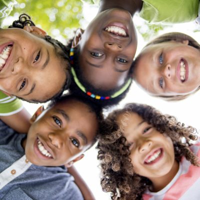 A multi-ethnic group of elementary age children are huddled together, standing in a circle with their heads in the center looking down at the camera. They are smiling and looking at the camera.